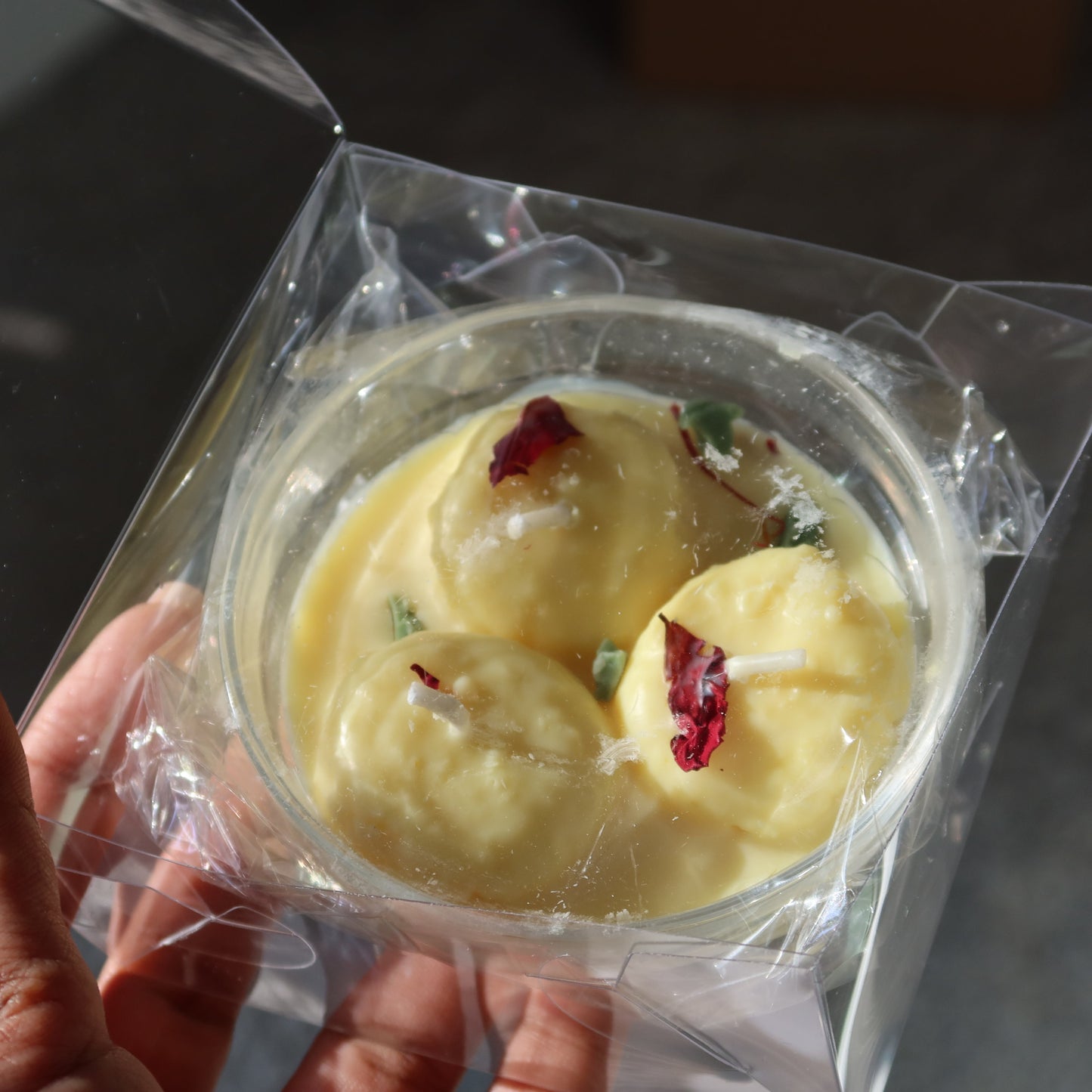 Hand holding a container of yellow dessert with dried fruits, wrapped in clear plastic.